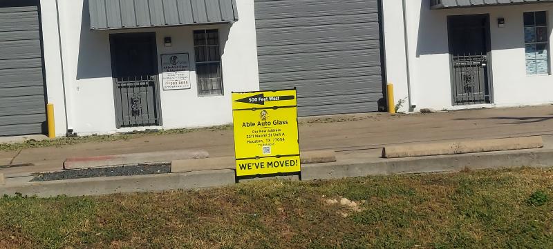 Able Auto Glass relocation sign with yellow background, black arrow pointing west labeled '500 feet west,' company name, new address at 2311 Naomi Street Unit A, Houston TX, QR code, and bold text reading 'WE'VE MOVED.